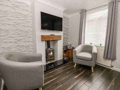 A living room with two grey armchairs a wood burning stove under a wall mounted tv and a window with blinds and curtains at Scarborough Cottage in Driffield