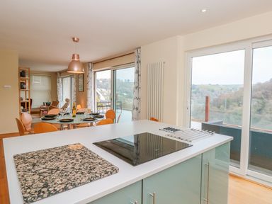 A kitchen island with a cooktop next to a dining table set with plates and glasses in a room with glass doors overlooking a valley at High Banks in Dartmouth