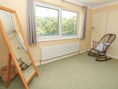 A room with a large window showing trees a wooden rocking chair with a cushion a full-length wooden framed mirror a floor lamp a basket and built-in wardrobes at High Banks in Dartmouth