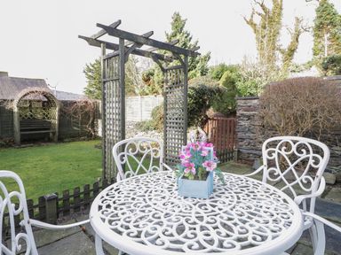 A garden with a white metal table and chairs with pink flowers on the table and a wooden archway in the background at Dalesway Cottage in Bowness-On-Windermere