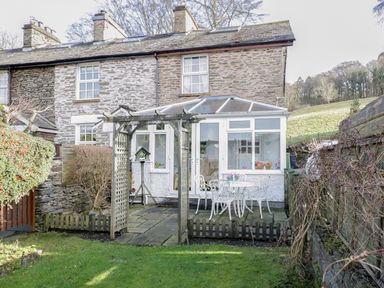 A backyard with a stone patio table and chairs and a wooden archway attached to a stone house at Dalesway Cottage in Bowness-On-Windermere