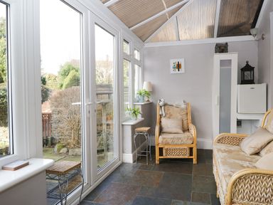 A room with wicker armchairs, a small white cabinet, a lamp on a side table, and large windows overlooking a garden at Dalesway Cottage in Bowness-On-Windermere