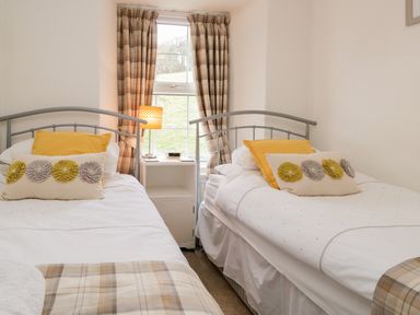 A bedroom with two single beds a nightstand lamp and plaid curtains at Dalesway Cottage in Bowness-On-Windermere
