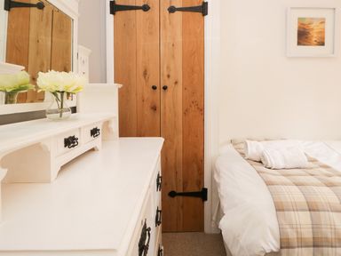 A bedroom with a white dresser with a mirror and flowers a wooden door and a bed with towels on a plaid blanket at Dalesway Cottage in Bowness-On-Windermere