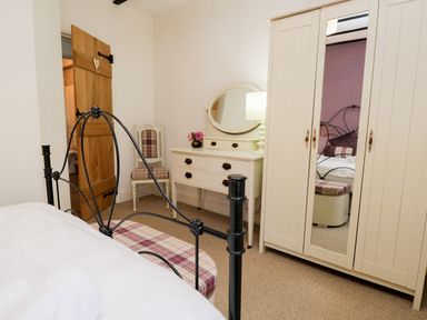 A bedroom with a black metal bed frame a white dresser with a mirror a chair with plaid upholstery and a wooden door at Dalesway Cottage in Bowness-On-Windermere
