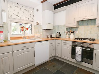 A kitchen with white cabinets a stove a dishwasher a sink and a window with a floral blind at Dalesway Cottage in Bowness-On-Windermere