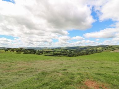 A green grassy field with trees and hills under a partly cloudy sky at Marches Way in Rhayader