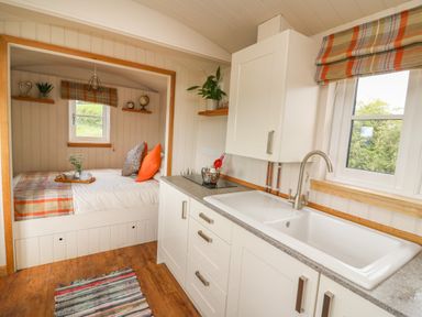 A small kitchen with white cabinets a sink and a window next to a built-in bed area with pillows and shelves at Marches Way in Rhayader