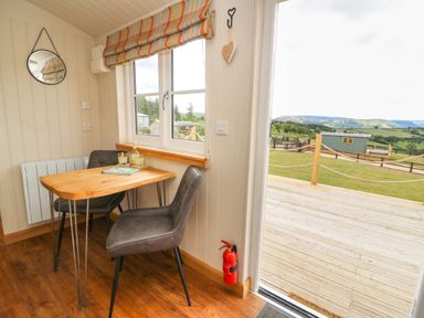 A small dining area with a wooden table and two chairs by a window with a striped blind and a door opening to a deck and grassy outdoor area at Marches Way in Rhayader