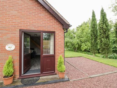 An open wooden door on a brick house showing part of the interior with potted plants outside and green trees at Byefield Lodge in Welland