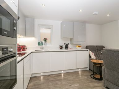 A kitchen with white cabinets a sink a microwave and oven a grey sofa and a wooden side table at Flat 2 38 Preston Road Weymouth