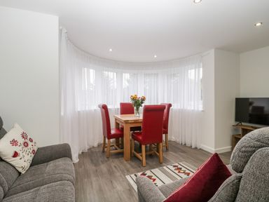 A dining area with a wooden table and red chairs next to curved windows with white curtains and a living space with grey sofas and a television at Flat 2 38 Preston Road in Weymouth