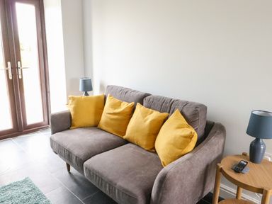 A gray sofa with four yellow cushions next to a small wooden table with a blue lamp at Tyddyn Haidd in Amlwch