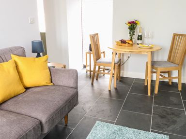 A living room with a gray sofa and yellow pillows next to a small wooden dining table with chairs and flowers at Tyddyn Haidd in Amlwch
