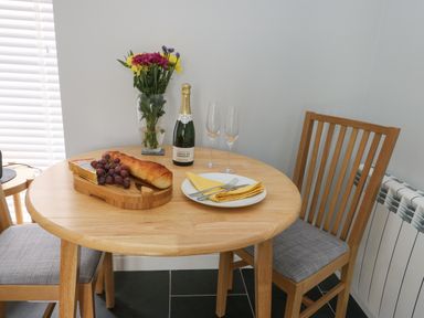 A wooden dining table with two chairs with a vase of flowers a bottle of champagne two champagne glasses a baguette grapes cheese and a plate with utensils at Tyddyn Haidd in Amlwch