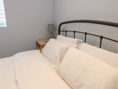 A bed with white bedding and pillows next to a wooden nightstand with a lamp at Tyddyn Haidd in Amlwch