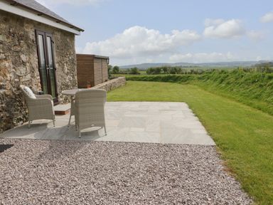 A stone building with a patio table and two chairs on a stone slab area next to a gravel path and lawn at Tyddyn Haidd in Amlwch