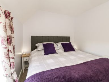 A bedroom with a double bed gray headboard purple cushions and blanket a bedside table with a lamp and floral curtains at Parva Great Broughton near Stokesley