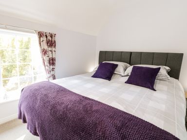 A bedroom with a double bed with purple pillows and blanket next to a window with floral curtains at Parva Great Broughton near Stokesley
