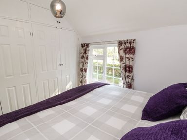 A bedroom with a bed covered in a checkered blanket and purple pillows next to a window with floral curtains and white built-in wardrobes at Parva Great Broughton near Stokesley