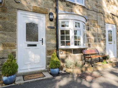 The exterior of a stone cottage with two white doors a bay window two potted plants and a wooden bench at Parva in Great Broughton near Stokesley