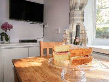 A kitchen with a wooden table holding a victoria sponge cake on a glass cake stand champagne bottle and two glasses a mounted TV and a potted plant on a cabinet near a window with checked curtains at Parva Great Broughton near Stokesley