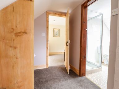 A hallway with two open doors one leading to a bathroom with a shower and toilet and the other to a carpeted room with framed wall art at Barn Court Cottage Narberth Pembrokeshire