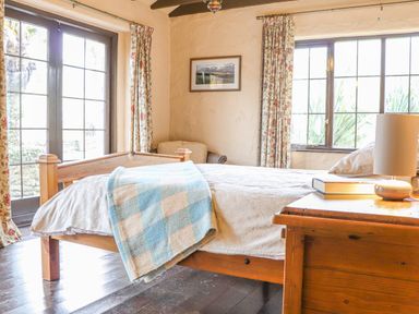 A bedroom with a wooden bed a blue and beige blanket floral curtains and a wooden bedside table with a lamp and books at Stoney Cottage in Narberth Pembrokeshire