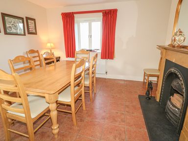 A dining room with a wooden table and six chairs a fireplace with a mirror and vase a window with red curtains and framed pictures on the wall at Goose Cottage in Caledon near Killylea