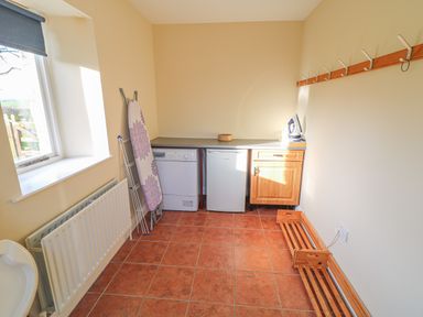 A utility room with a dishwasher and mini fridge under a counter with an iron on top an ironing board and drying rack near a window with wall hooks and wooden shelves on the side at Goose Cottage Caledon near Killylea