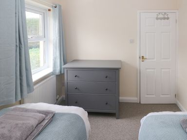 A bedroom with two single beds a gray chest of drawers a window with curtains and a white door at Smugglers in Lydstep