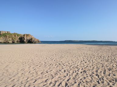 A sandy beach with footprints and a rocky cliff on the left overlooking the sea under a clear sky at Smugglers in Lydstep
