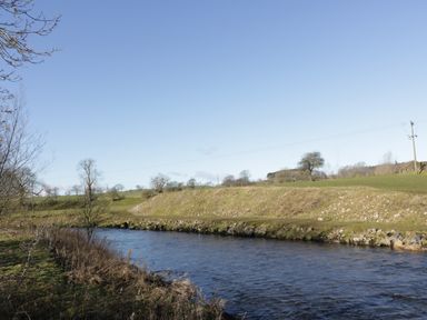 A river flowing through a grassy landscape with trees and a clear sky at Wren Lodge in Gargrave