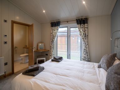 A bedroom with a bed with folded towels a small corner desk with a mirror and a stool open door to a bathroom and glass doors with curtains at Wren Lodge in Gargrave