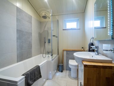 A bathroom with a bathtub shower combination a toilet a sink with a wooden cabinet and a small window at Wren Lodge in Gargrave