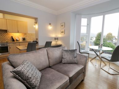 A living area with a grey sofa pillows a glass dining table with two chairs and a kitchen in the background at Tethera in Bowness-On-Windermere