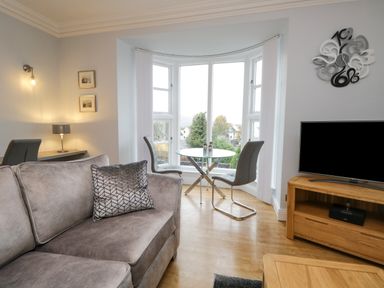 A living room with a grey sofa a glass dining table two chairs a wooden tv stand and a modern clock at Tethera in Bowness-On-Windermere