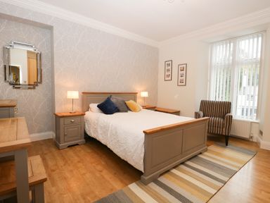 A bedroom with a bed with pillows two bedside tables with lamps an armchair beside a large window and a rug on the floor at Tethera in Bowness-On-Windermere