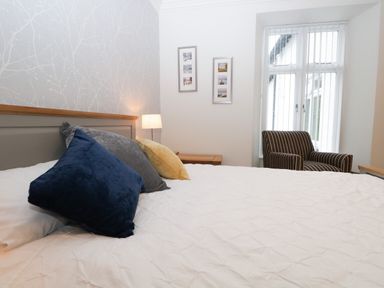 A bedroom with a bed with blue gray and yellow pillows a bedside table with a lamp two framed pictures on the wall and a striped armchair near a window at Tethera in Bowness-On-Windermere