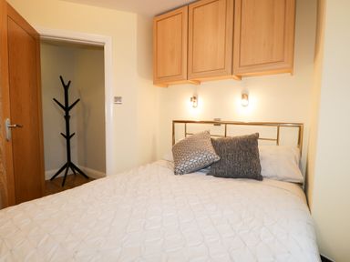 A bedroom with a bed with two pillows and a quilt under wooden cabinets near a wooden door with a coat rack visible at Tethera in Bowness-On-Windermere