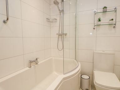 A bathroom with a bathtub shower combination a toilet a small trash bin and a glass shelf with plants at Tethera in Bowness-On-Windermere