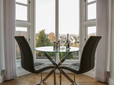 A dining area with a round glass table and two chairs by large windows showing an outdoor view at Tethera in Bowness-On-Windermere