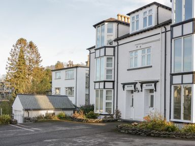 The exterior of a white multi-story building with large windows a small garage and a parking area surrounded by trees at Tethera in Bowness-On-Windermere