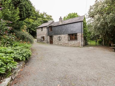 A gravel driveway leading to a stone house with a slate roof surrounded by trees and plants at Ribby Barn in Lerryn