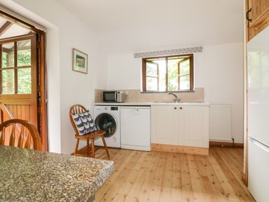 A kitchen with wooden floor and white cabinets a microwave a washing machine a dishwasher a wooden chair a window and a wooden door at Ribby Barn in Lerryn