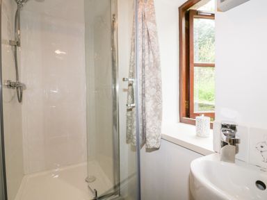 A bathroom with a glass shower enclosure a small sink a window with a floral curtain and a soap dispenser on the windowsill at Ribby Barn in Lerryn
