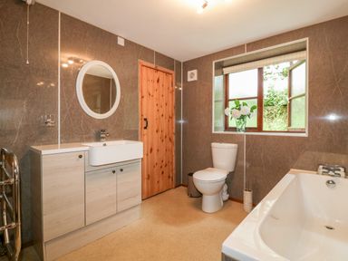 A bathroom with a wooden door a white sink with cabinet a round mirror a toilet a bathtub and a window with flowers on the sill at Ribby Barn in Lerryn