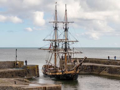 A tall ship with two masts docked in a stone harbor with people standing on the harbor walls and the sea in the background at Ribby Barn in Lerryn