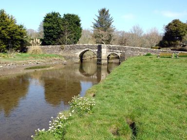 A stone bridge over a shallow river with grassy banks and trees in the background at Ribby Barn in Lerryn