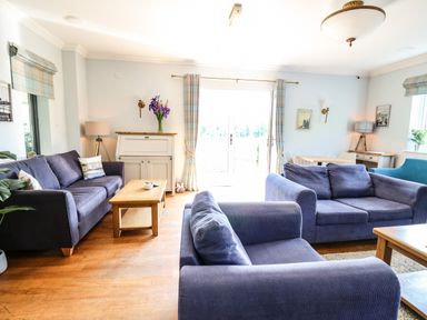 A living room with blue sofas wooden coffee tables a white cabinet with flowers and large windows at Llety Ceiro in Bow Street Wales